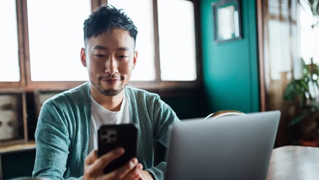 Man holding and looking at a smartphone and sitting in front of laptop