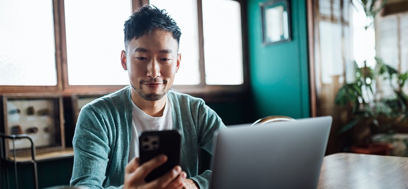 Man holding and looking at a smartphone and sitting in front of laptop